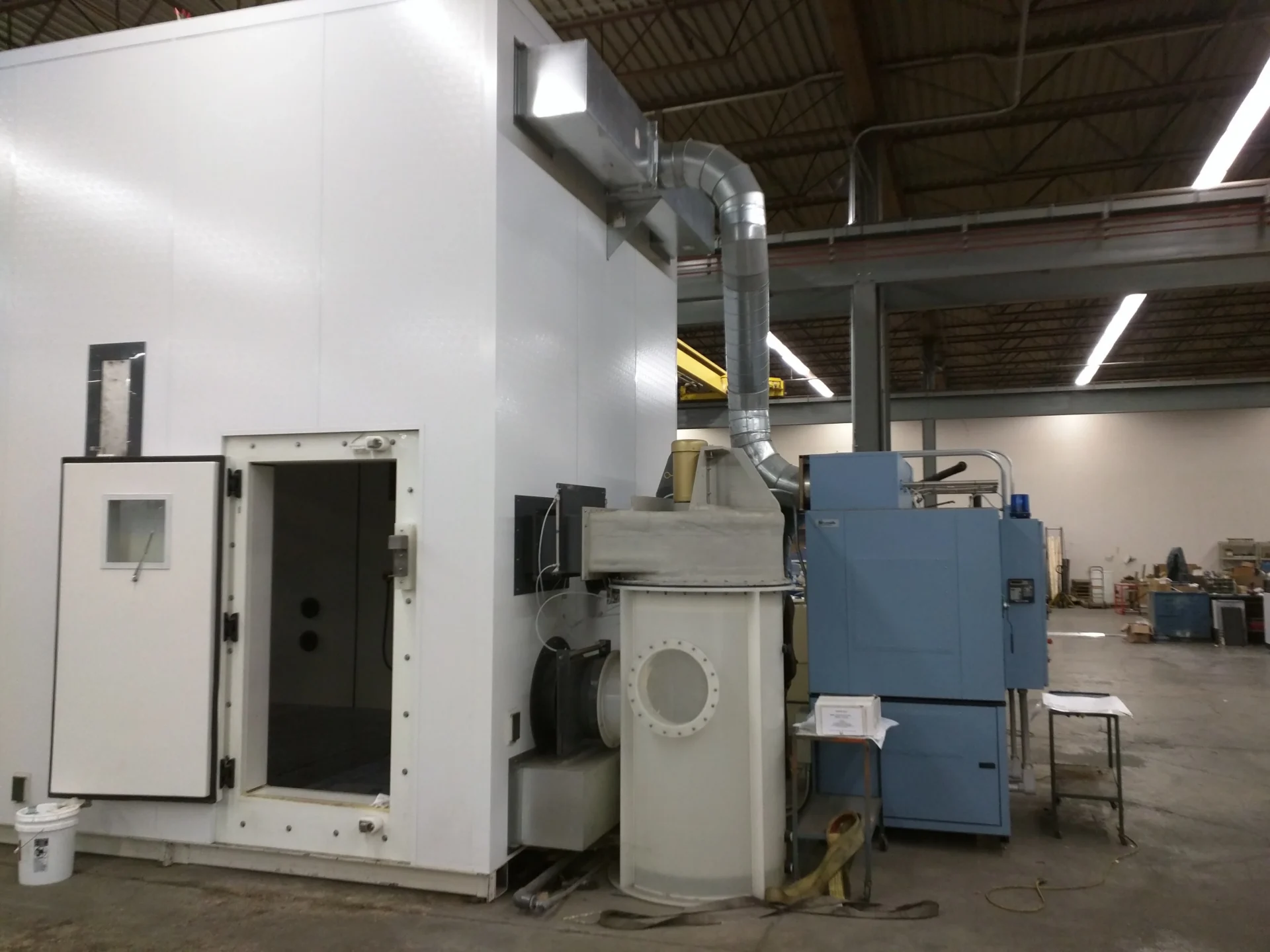 Large white walk-in industrial corrosion test chamber with an open heavy-duty door, connected to a vertical exhaust scrubber tank and a blue mechanical cooling refrigeration unit in a warehouse facility.