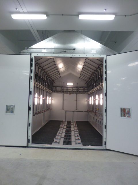 Interior of a 13 by 25 foot drive-in corrosion chamber showing complex wall-mounted, overhead, and floor-level spray nozzle manifolds for uniform sample coverage.