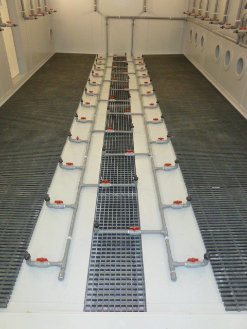 Close-up of floor-mounted gray PVC piping with individual control valves and spray nozzles situated between non-slip floor grates inside a test chamber.