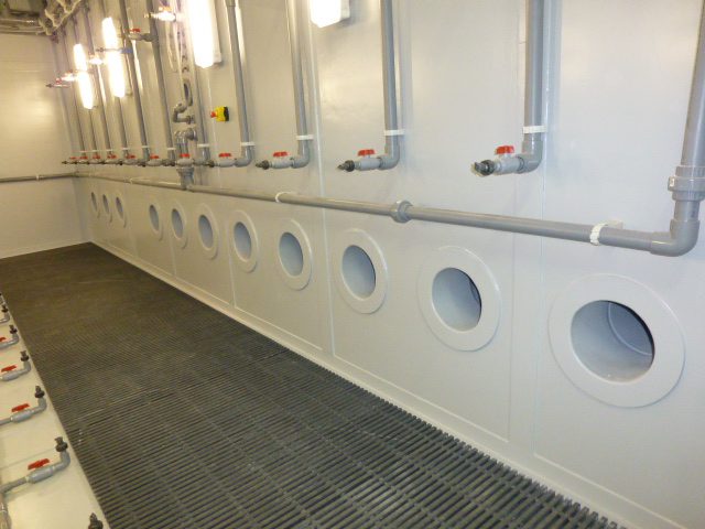 Side wall view inside an environmental chamber showing a long row of large circular air ports located beneath direct spray pipes and industrial lights.