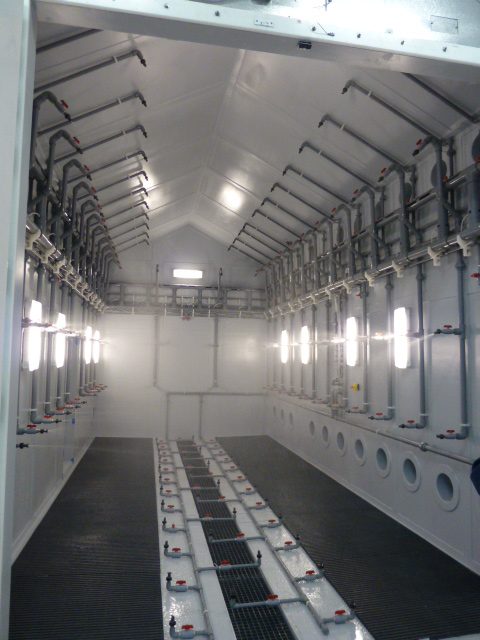 Looking through the wide double doors into an empty white corrosion chamber equipped with extensive gray PVC fluid piping and arrayed side wall lighting.