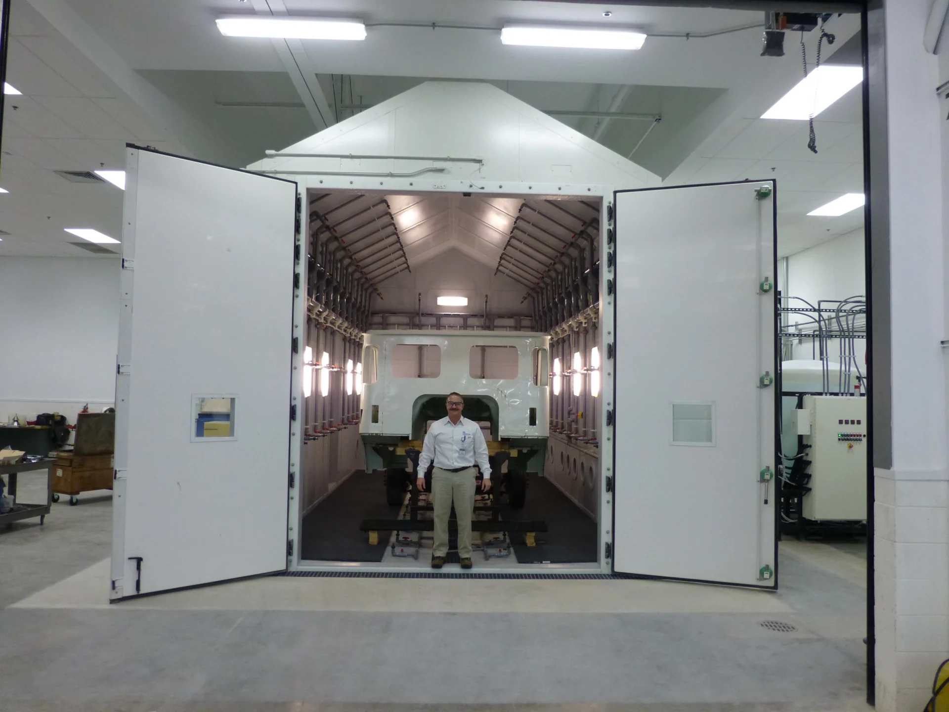 A technician standing in front of an open drive-in corrosion chamber containing a large commercial vehicle cab section for structural spray testing.