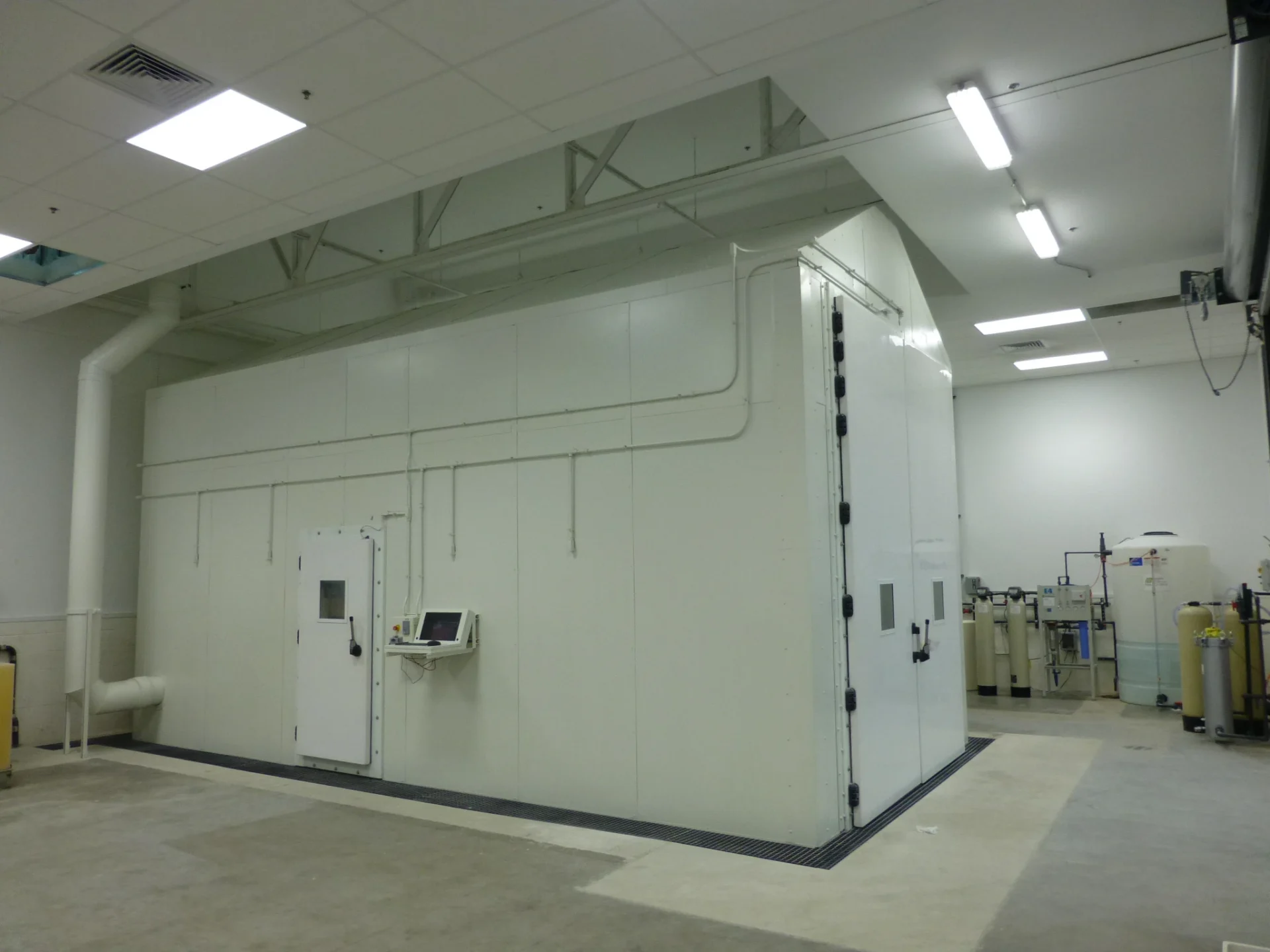 Modular exterior paneling of a large white drive-in environmental test chamber with an external access door, a localized operator monitor, and water filtration tanks nearby.